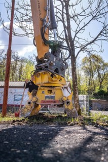 An excavator with grab arm stands in front of a bare tree and a building, demolition site,