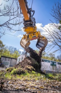 An excavator with a grab arm lifts earth while the sun is shining and trees and buildings can be