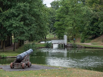 A historic cannon stands in a park on the banks of a river, surrounded by green trees and a small