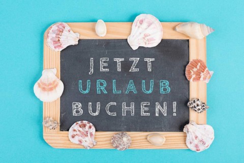 Book vacation now is standing in german language on the chalkboard, seashells bulit frame, holiday