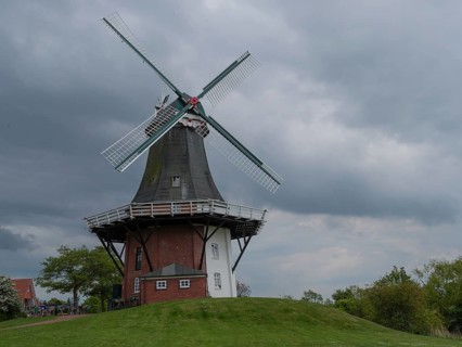 Old windmill on a green hill in front of a cloudy sky and a brick white building, greetsiel, east