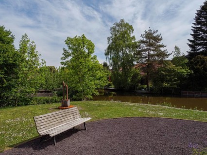A park bench stands on a green meadow next to a small fountain, at the edge of a river with trees,