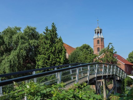 Wooden bridge leading to a church tower, surrounded by lush green vegetation and blue sky, Ditzum,