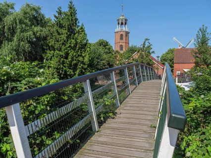 Wooden bridge leads to a church tower and a windmill, surrounded by trees under a blue sky, Ditzum,