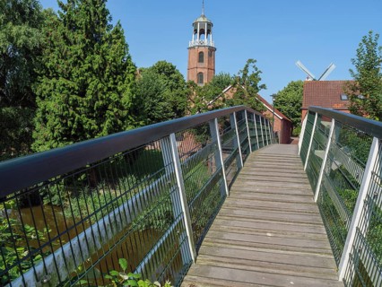 Wooden footbridge leading to a historic church tower, surrounded by green vegetation and clear