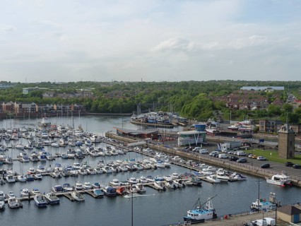 A harbour full of boats and yachts, surrounded by green spaces and houses under a cloudy sky,