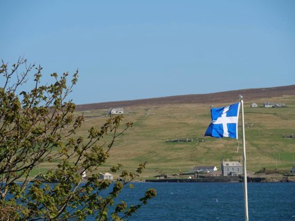 A Scottish flag flies on the coast with green hills and scattered houses in the background,
