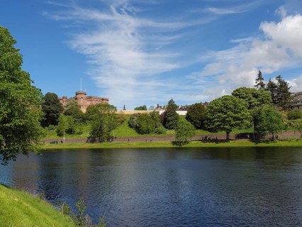 A river flows in front of a green hill and a castle, surrounded by lush vegetation and a clear sky,