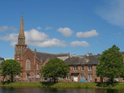 Church tower and urban buildings along a river with trees and blue sky with clouds, inverness,
