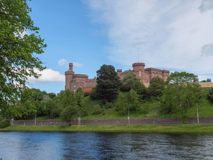 A castle overlooking a river, surrounded by green banks and trees under a blue sky, inverness,