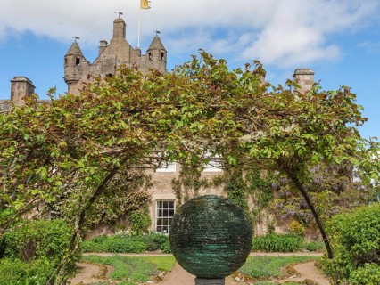 Castle with towers in the background, an archway of plants and a sculpture in a well-tended garden,