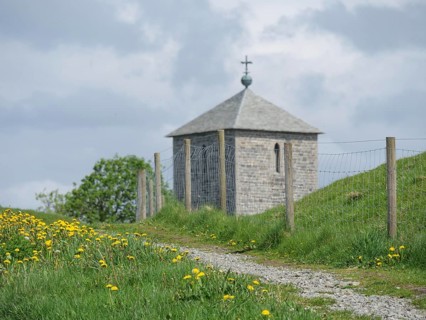 Stone church with picket fence on a green path, surrounded by yellow flowers and cloudy sky,