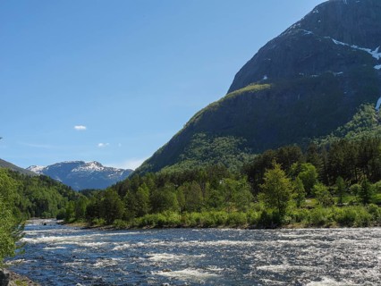 A river flowing through a green, forested landscape, surrounded by mountains under a clear sky,
