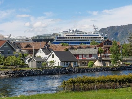 A riverside village with a large cruise ship in the background, surrounded by mountains under a