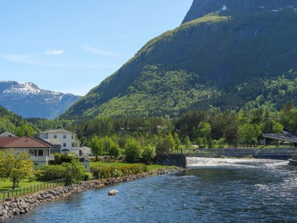 A river with a bridge, flanked by houses and trees, embedded in a green valley with mountains in