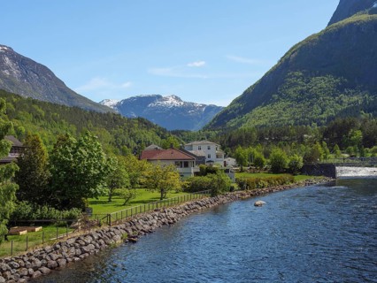 A river flows past houses and trees in a green valley surrounded by high mountains, Eidfjörd,