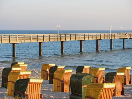Colourful beach chairs stand on the sandy beach in front of a long wooden jetty over the calm sea,