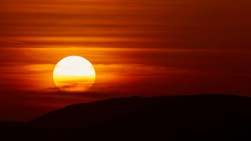 The setting sun breaks through dramatic layers of cloud and illuminates the silhouette of a hill,