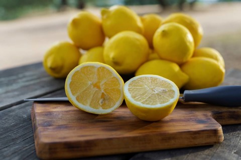 Lemon cut with a knife on a wooden board on a table in a park with a pile of lemons and trees in