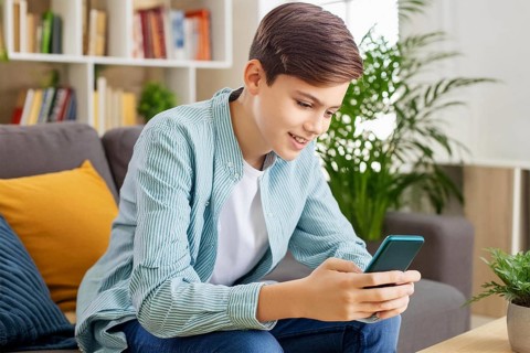 A smiling boy in a striped shirt sits on a sofa and uses his smartphone, AI generated, AI generated