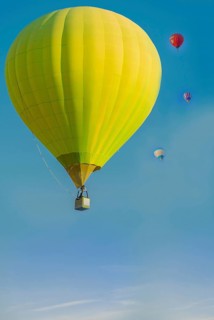 A bright yellow hot air balloon floating in a clear blue sky with smaller colorful balloons in the