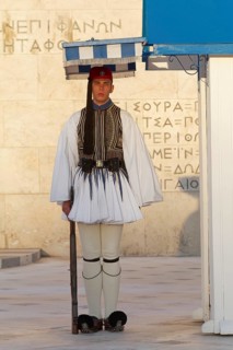 Evzone in traditional dress stands next to a guard hut under a sunshade, Parliament building, Tomb