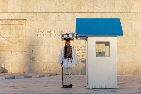 Evzone in traditional dress stands next to a guard hut under a sunshade, Parliament building, Tomb