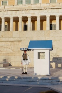 Evzone in traditional dress stands next to a guard hut under a sunshade, Parliament building, Tomb