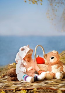 A baby dressed in a pink outfit sits on a blanket near a lake, accompanied by a teddy bear and a