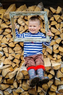 A smiling boy in a striped shirt and boots sits on a woodpile, holding a rustic, weathered frame