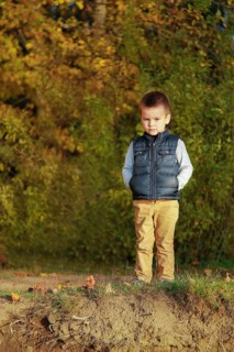 A young boy in a blue vest stands on a grassy area surrounded by autumn leaves and trees, Belarus