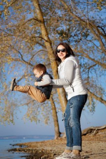 A woman lifting a young boy near a lake on a sunny autumn day with trees and blue sky in the