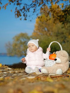 A baby sitting on a blanket outdoors in autumn with a teddy bear, a basket containing a pumpkin and