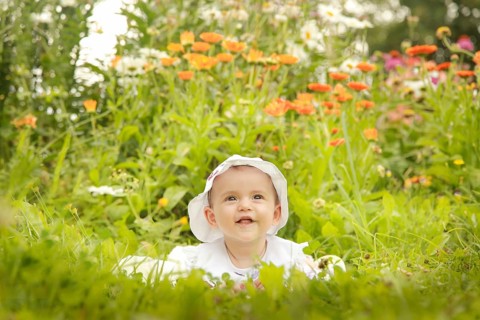A smiling baby wearing a white hat sits amidst a garden filled with colorful flowers and lush