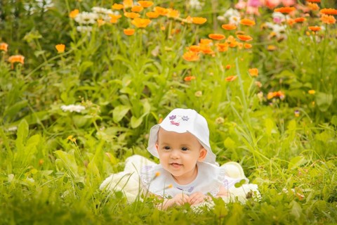 A baby wearing a hat lying in the grass surrounded by colorful flowers on a sunny day, Belarus