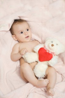 A baby lying on a pink blanket holding a white teddy bear with a red heart