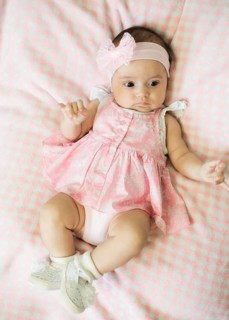 Baby in a pink dress with a headband and bow, lying down on checkered bedding, wearing white shoes
