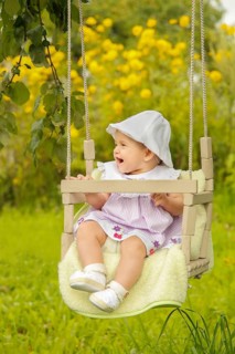 A baby sitting on a swing in a lush garden with yellow flowers, wearing a hat and dress while