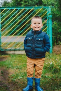 Young boy wearing a blue jacket and boots stands outdoors near a green gate.Belarus.Minsk