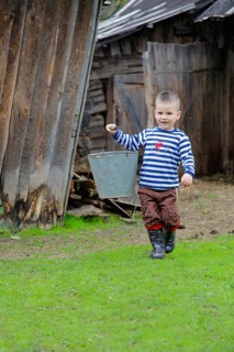 A boy carrying a metal bucket, wearing a striped shirt, brown pants, and rubber boots, walking on
