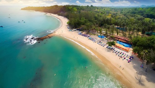 A beautiful beach with a clear blue ocean and a green forest in the background. The beach is full