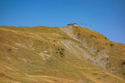 Extensive mountain landscape with a hut on a hill, crossed by a cable car under a blue sky,