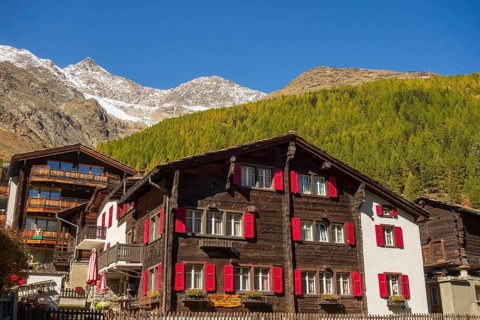A traditional house with red shutters against a backdrop of forest and mountains in autumn, Saas