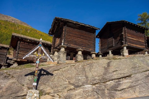Traditional wooden houses and a cross on a rocky mountainside under a blue sky, Saas Fee,