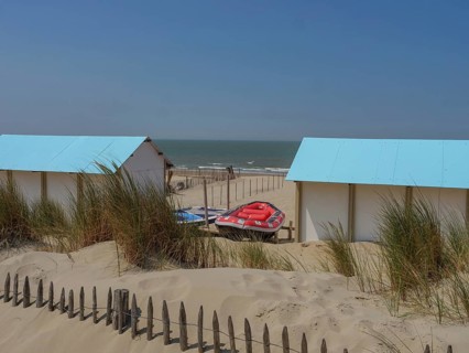 Wooden huts on the beach, surrounded by sand dunes and lifeboats, with the sea in the background,