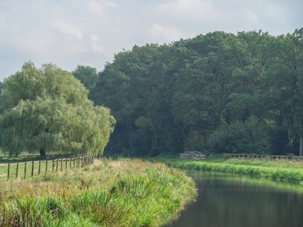 A calm river flows along a wooded bank and a green meadow under a cloudy sky, haaksbergen, .