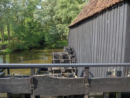 A historic wooden building next to a river and a bridge, surrounded by dense forest, haaksbergen, .