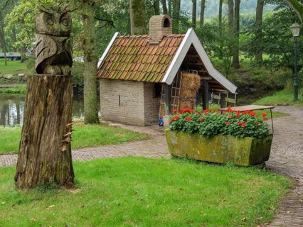 A rural garden with a carved wooden owl, a small brick house, a profusion of flowers and old trees,