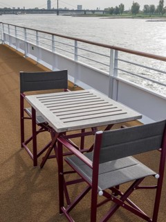 Deck of a ship with table and chairs, view of the river and the surrounding landscape. The railing