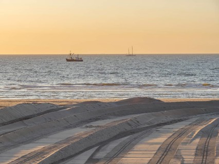 A sunset over the sea with silhouettes of boats and a sandy beach in the foreground, de haan,